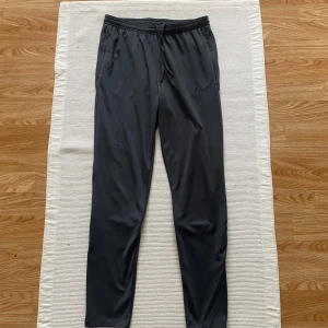 Grå Nike Dri-Fit träningsbyxor - Grå träningsbyxor från Nike med Dri-Fit-teknologi, elastisk midja med snörning och diskret logga på benet. Byxorna har smal passform, dragkedjeförsedda fickor och är gjorda i lätt polyester som andas. Perfekta för sport och aktivitet.