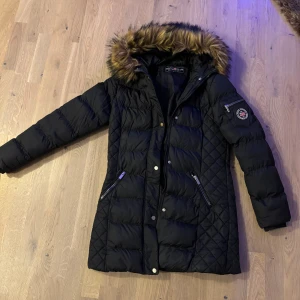 Svart lång pufferjacka med päls - Snygg svart pufferjacka med quiltad design och fuskpäls på huvan. Jackan har dragkedjor på fickorna, tryckknappar framtill och ett märke på ärmen. Perfekt för kalla dagar och har en längre passform.