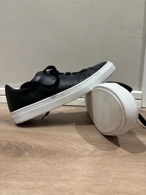 Svarta sneakers från Björn Borg - Snygga svarta sneakers från Björn Borg med vit platt sula. Skorna har snörning och är tillverkade i skinn med en stilren och klassisk design. Perfekta för dig som gillar minimalistisk stil och vill ha ett par schysstare sneakers till vardags.              Mycket bra skick men lite creasade där fram. Använde dem endast under en helg då de var lite små för mig.