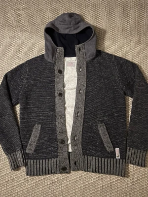 Mörkgrå stickad cardigan jacka Replay - Säljer en mörkgrå stickad cardigan jacka från Replay med kontrasterande ljusgrå ribbade detaljer och huva i sweatshirtmaterial. Jackan har knappar framtill, två fickor och ribbade muddar. Perfekt för dig som gillar en chill och avslappnad stil. Den saknar en knapp i mitten