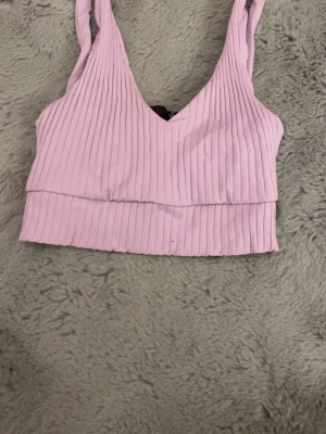 Ljuslila ribbad linnetopp - Säljer en ljuslila croppad linnetopp med ribbad struktur och v-ringad hals. Toppen är ärmlös och har breda axelband, perfekt för varma dagar. Materialet känns mjukt och stretchigt mot huden. Kan eventuellt tänka mig gå ner i pris