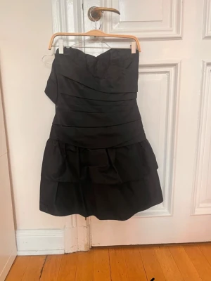 Svart axelbandslös kortklänning - Snygg svart axelbandslös klänning med volangdetaljer och omlottliknande lager på överdelen. Klänningen har en figurnära passform upptill och en fluffig kjol med flera lager. Dragkedja baktill för enkel påklädning. Perfekt för dig som gillar stilrena och trendiga plagg.