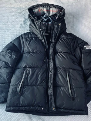 Svart pufferjacka från Burberry - Svart pufferjacka från Burberry med huva och dragkedja. Jackan har ett rutigt foder i grått, vitt och rött, samt Burberry London England-logga på ärmen. Två fickor med dragkedja framtill och quiltad design. Perfekt för kalla dagar.