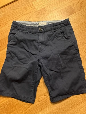 Mörkblå shorts från Lindex - Snygga mörkblå shorts från Lindex med klassisk passform och midwaist. De har knapp och dragkedja framtill, samt praktiska fickor på sidorna. Perfekta för varma dagar och enkla att matcha med t-shirt eller skjorta. Materialet är mjuk bomull som känns skönt mot huden.
