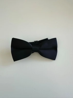 Svart fluga i satin med justerbart band - Snygg svart fluga med klassisk form och justerbart band. Flugan är tillverkad i slät satin som ger en stilren look. Perfekt accessoar för att lyfta din outfit till nästa nivå. Levereras helt ny i plastförpackning.