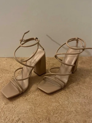 Beige sandaler med blockklack - Snygga beige sandaler med höga blockklackar och smala remmar som slingrar sig runt foten och ankeln. Sandalerna har öppen tå och fyrkantig sula, vilket ger en modern vibe. Perfekta för dig som gillar stilrena och trendiga skor med extra höjd.