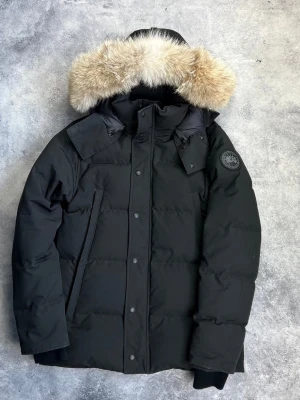 Svart Canada Goose Label dunjacka med päls - Säljer en svart dunjacka från Canada Goose med fluffig päls på huvan. Jackan har lång ärm, dragkedja och knappar framtill, samt märkets patch på ärmen. Perfekt för kalla vinterdagar och har en clean, stilren look.