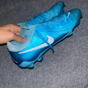 Nike blå fotbollsskor Flyknit - Säljer ett par blå Nike fotbollsskor med vit swoosh och snörning. Skorna har Flyknit-material för extra komfort och passform, samt platt sula med dobbar för gräsplan. Snygg design med ljusblå och turkosa toner, perfekt för dig som vill sticka ut på planen.
