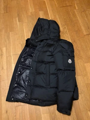 Svart Moncler dunjacka med huva - Svart dunjacka från Moncler med stor huva och quiltad design. Jackan har dragkedja och tryckknappar framtill, samt en ficka med Moncler-logga på ärmen. Insidan har ett coolt serietidningsmönster och flera etiketter. Perfekt för kalla dagar. Pris kan diskuteras vid snabbaffär