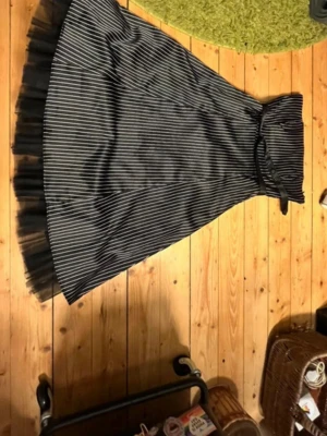 Svart axelbandslös randig långklänning - Snygg svart axelbandslös långklänning med vita ränder och ett brett bälte i midjan. Klänningen har volangkant i tyll nedtill och en omlottliknande överdel. Materialet är glansigt och känns som polyester. Perfekt för dig som gillar en cool och elegant look.