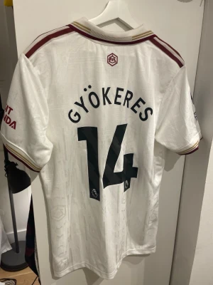 Arsenal Gyökeres Fotbollströja - Säljer en vit Arsenal fotbollströja från Adidas i storlek XL med Gyökeres och nummer 14 på ryggen. Tröjan har vinröda och guldiga detaljer på krage och ärmar, samt broderat klubbmärke och Adidas-logga framtill. Materialet är syntet och designen är inspirerad av klassiska retrotröjor. Säljer den pga att jag inte har någon användning för den