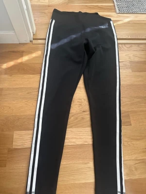 DESIGN 2 MOVE 3-STRIPES HIGH-RISE LONG TIGHTS - Svarta Adidas tights med tre vita ränder längs benen. Tightsen har en Adidas logga fram och Aeroready logga bak. Mycket fint skick förutom att sömmen har släppt något vid ena randen (inget som stör). Kan enkelt sys tillbaka om man är händig