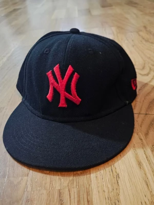 Svart New Era keps med röd NY-logga - Säljer en svart New Era 59FIFTY keps med röd broderad NY-logga framtill och röd MLB-logga bak. Klassisk flat peak och stilren look, perfekt för dig som gillar streetwear. Kepsen är tillverkad i bomull och har äkta merchandise-märkning.