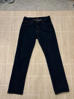 Mörkblå Lee jeans W31 L32 - Säljer ett par mörkblå jeans från Lee i modellen West. Klassisk femficksdesign med raka ben och normal passform. Snygga kontrastsömmar och metallknapp med Lee-logga. Jeansen har blivit använda ungefär 10 gånger och är i ett bra skick. Killen i bilden är 172 cm för referens.