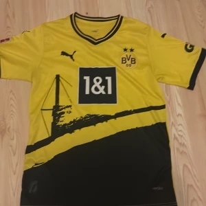 BVB Dortmund fotbollströja Puma gul - Säljer en officiell Borussia Dortmund fotbollströja från Puma i storlek S. Tröjan är gul med svarta detaljer, har BVB 09-emblem, Bundesliga-märke på ärmen och sponsortryck 1&1 på bröstet. Snyggt grafiskt tryck framtill och DORTMUND på ryggen.