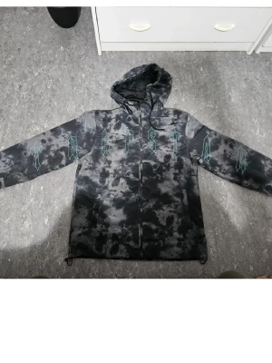 Svart och grå vindjacka från Trapstar  - Säljer en svart och grå tie-dye vindjacka från Trapstar med huva och dragkedja framtill. Jackan har långa ärmar och coolt tryck i turkos på bröstet och armarna. Perfekt för dig som gillar streetwear och vill sticka ut med en unik look.