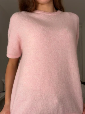 Ljusrosa stickad topp med kort ärm - Supermjuk ljusrosa stickad topp med rund hals och korta ärmar. Enkel och clean design utan mönster eller detaljer, perfekt att styla med jeans eller kjol. Materialet är fluffigt och känns skönt mot huden.