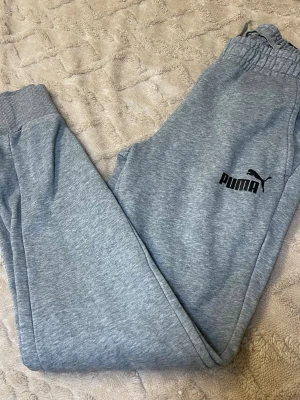 Grå mjukisbyxor från Puma - Snygga grå mjukisbyxor från Puma med svart logga på vänster ben. Byxorna har elastisk midja med snörning och ribbade muddar vid bensluten. Tillverkade i mjukt bomullsmaterial, perfekta för chill eller träning. Fickor på sidorna för extra funktion.