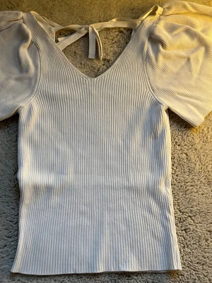 Vit ribbad topp med puffärm från ONLY - Säljer en vit ribbad topp från ONLY i storlek S. Toppen har korta puffärmar, v-ringning och snygg knytning i nacken. Materialet är mjukt och stretchigt, perfekt för dig som gillar en feminin och trendig stil.