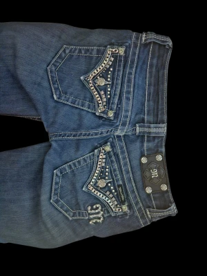 Blå Miss Me jeans - Säljer ett par blå jeans från Miss Me med snygga kedjedetaljer och nitar på fickorna både fram och bak. Jeansen har kontrastsömmar, låg midja och bootcut-ben. Märkeslogga på bakfickan och metallknappar. 