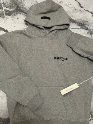 Essentials hoodie grå – Helt Ny S - Helt ny Essentials Fear of God hoodie i storlek S. Aldrig använd, med tag kvar.  Mjuk och skön passform. Perfekt för vardag och streetwear 🔥✨ Pusha och köp snabbt ⏳💨 – köp innan någon annan gör det! 🛒
