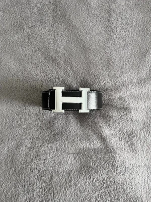 Hermés bälte  - Säljer ett svart skärp från Hermès med ett stort H-format metallspänne i silverfärg. Skärpet är gjort i äkta skinn med tydliga vita sömmar längs kanterna och har en stilren, klassisk look. Perfekt för att lyfta din outfit med en lyxig detalj.