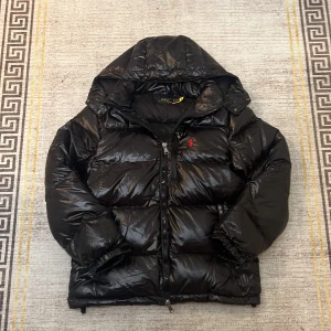 Svart pufferjacka från Polo Ralph Lauren - Svart pufferjacka med glansig finish från Polo Ralph Lauren. Jackan har huva, dragkedja framtill och röd broderad logga på bröstet. Fylld med syntetmaterial och har elastiska muddar vid ärmslut. Perfekt för kalla dagar och riktigt snygg streetstil.