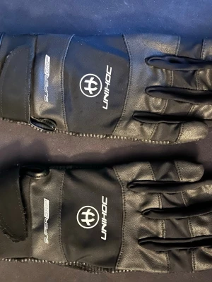 Svarta Unihoc SuperGrip handskar| helt nya köpta från klubbhusets butik aldrig använda.  - Säljer ett par svarta Unihoc SuperGrip handskar med snyggt diagonalt mönster på insidan för extra grepp. Handskarna har justerbar kardborre vid handleden och är tillverkade i syntetmaterial med detaljer i mesh och gummi. Perfekt för innebandy eller annan sport.