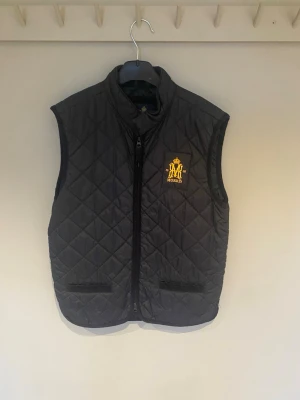 Svart quiltad väst från Morris - Quiltad svart väst från Morris med gul broderad logga på bröstet. Västen har ståkrage, två fickor framtill och dragkedja. Materialet är polyester och passformen är normal. Perfekt för lager-på-lager under höst och vinter.