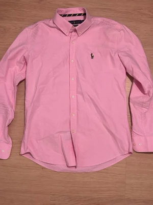 Rosa skjorta från Ralph Lauren - Klassisk rosa skjorta från Ralph Lauren med button-down krage och broderad logga på bröstet. Skjortan har långa ärmar, vita knappar och är tillverkad i mjuk bomull. Perfekt för dig som vill ha en stilren och tidlös look. Köpt här på plick. Passar storlek S.