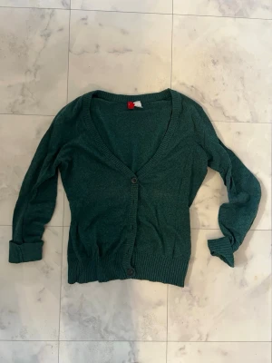 Grön kofta från H&M Divided - Mörkgrön kofta från H&M Divided i storlek 34. Koftan har v-ringad hals, långa ärmar och knäppning med två knappar framtill. Ribbstickade muddar vid ärmslut och nederkant ger en snygg och bekväm passform. Perfekt att slänga på över en t-shirt.