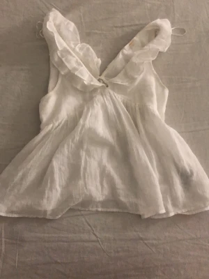 Vit blus med volang och omlott - Säljer en söt vit blus i tunn och luftig bomull med omlott framtill och breda volangdetaljer vid axlarna. Blusen är ärmlös och har en lätt, flowy passform som ger en romantisk vibe. Perfekt för varma dagar och sommarhäng.