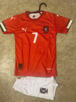 Portugisiska fotbollsshorts och tröja - Säljer ett set med Portugals landslagströja och shorts. Tröjan är röd med vita detaljer, nummer 7 och landslagsemblem, samt 'Ronaldo' på ryggen. Shorts är vita med emblem. Materialet är polyester och har DryCell-teknologi för att hålla dig torr.