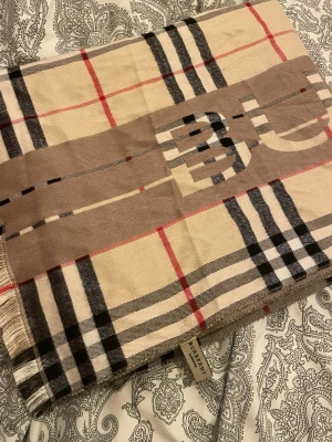 Beige rutig halsduk - Säljer en beige halsduk med klassiskt rutigt mönster i svart, rött och vitt. Perfekt för dig som vill ha en ikonisk och stilren look med lyxig touch.