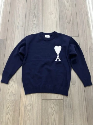 Mörkblå stickad tröja från Ami - Snygg mörkblå stickad tröja från Ami med ett vitt hjärta och bokstaven A broderat på bröstet. Tröjan har rund hals, ribbade muddar och långärmad passform. Perfekt för dig som gillar stilrena detaljer och vill ha något som sticker ut lite.