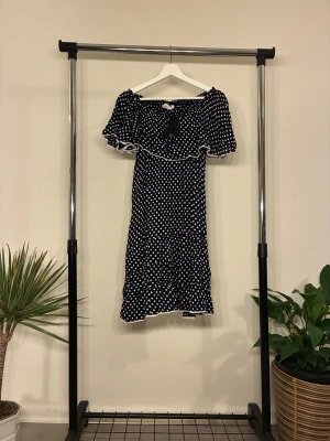 Prickig klänning  - Supersöt svart klänning från Isolde, Offshoulder-modell med volang upptill och vita prickar över hela. Klänningen har kort ärm och avslutas med en liten spetskant. Perfekt för dig som gillar en lekfull och trendig look. OANVÄND 