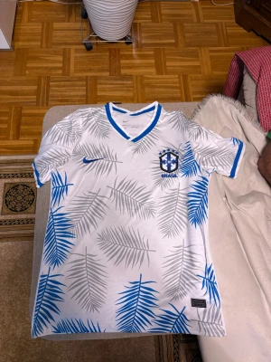 Brasilien fotbollströja - Jag säljer en vit och blå Brasilien Nike fotbollströja med palmbladsmönster i storlek S men passar mer som M. Den är oanvänd och säljs då den var lite för stor för mig.