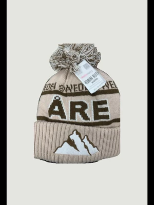 Åre mössa - Bilden visar en beige och brun stickad mössa med texten ÅRE och ett bergmotiv, samt en stor tofs på toppen. Ingen väst syns på bilden. Skriv vid minsta lila fråga. Pris ej hugget i sten 🪨 