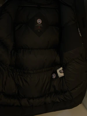 Svart dunjacka från Canada Goose - Säljer en svart dunjacka från Canada Goose med huva och klassisk logga på ärmen. Jackan har dragkedja framtill, stora fickor och är perfekt för kalla dagar. Materialet är slitstark polyester och dunfyllning som håller dig varm hela vintern.