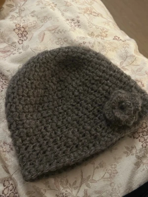 Handgjord mössa med blomma - Mysig grå virkad mössa med en blomma på sidan. Mössan är handgjord med mohair och ull blandning garn som gör den mjuk och håller dig varm.  Pga av materialet är den lite stretchig därför kan man inte utgå ifrån måtten helt. Längd: 20 cm   omkrets: 50 cm