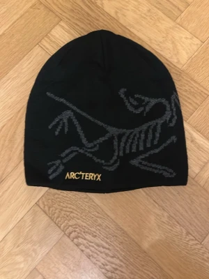 Svart mössa från Arc'teryx - Svart mössa från Arc'teryx med grått skelettmönster på framsidan och gul broderad logga. Snygg och enkel design som passar perfekt till kalla dagar. Materialet känns mjukt och stretchigt, vilket gör den bekväm att bära.