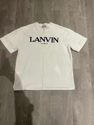 Vit Lanvin Paris t-shirt med broderi - Vit t-shirt från Lanvin Paris med svart broderad logga framtill och ett diskret vitt broderi i nacken. Klassisk rund hals och korta ärmar. Tillverkad i mjuk bomull som känns skön mot huden. Perfekt för dig som gillar stilrena designer och exklusiva märken.