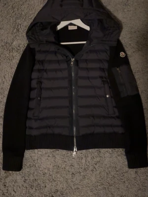 Svart pufferjacka från Moncler - Säljer en svart pufferjacka från Moncler med huva och ribbstickade ärmar. Jackan har två sidofickor med tryckknappar, dragkedja framtill och en extra ficka med dragkedja på ena ärmen. Materialmix av polyester och stickat ger en schysst kontrast.  Storlek M passar S jackan har inga skador