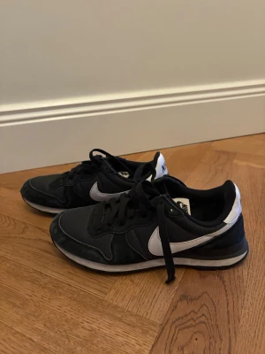 Svarta Nike sneakers med vita detaljer  - Jättesnygga sneakers ifrån märket Nike i svart med vita detaljer och vit häl. Skorna har snörning, rund tå och platt sula. Ovandelen är i mesh och mocka, vilket ger en skön och sportig känsla. De är klassiska, sköna och i bra material. De är i okej skick men lite slitna med små små fläckar som knappt syns, därför priset. De är i storlek 38 och ett perfekt basplag som passar allt och som är perfekt nu till våren. Skriv om funderingar, lämna gärna omdöme och priset kan diskuteras 😊
