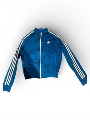Blå vintage Adidas kofta - Snygg vintage kofta från Adidas som inte säljs längre  med klassiska vita ränder längs ärmarna och vit dragkedja. Koftan har ribbade muddar, lång ärm och Adidas-logga på bröstet. Perfekt för dig som gillar sportig stil och streetwear.