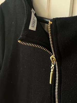 Svart klänning från Soaked in Luxury - Stilren svart klänning från Soaked in Luxury i storlek S. Rak och kort passform, trekvartsärm och en snygg guldfärgad dragkedja i ryggen. Tidlös minimalistisk design. Kan stylas med höftskärp som variation och snyggt med statementhalsband i halsringningen. 