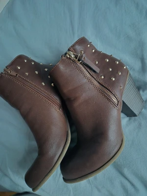 Bruna boots med nitar och klack - Snygga bruna boots med grov klack och rund tå. Skorna har dekorativa guldfärgade nitar på skaftet och dragkedja på sidan för enkel på- och avtagning. Tillverkade i skinnimitation med en stabil plattformsula. Nästqn helt nya använda fåtal gånger.💕