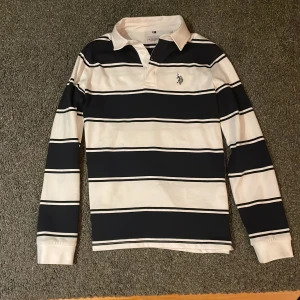 Pikétröja från U.S. Polo Assn. - Säljer en stilren randig pikétröja från U.S. Polo Assn. i marinblått och vitt. Tröjan har långa ärmar och en klassisk krage med knappar. Den är aldrig använd helt ny skick. Köpt för 499.