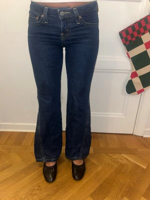 Levi's Noughties Bootcut Jeans mörkblå - Säljer ett par mörkblå Levi's Noughties Bootcut jeans i storlek 25. Klassisk femficksmodell med kontrastsömmar, låg midja och bootcut-ben. Jeansen har snygga detaljer på bakfickorna och är tillverkade i slitstarkt denimtyg