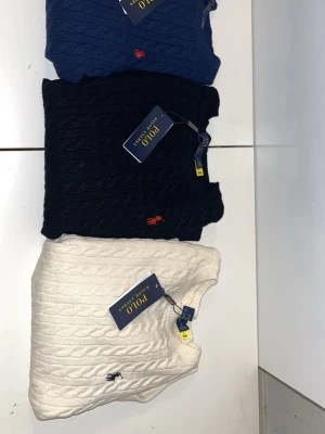 Kabelstickad tröja Polo Ralph Lauren - Kabelstickad tröja från Polo Ralph Lauren i tre färger: marinblå, svart och vit. Tröjan har rund hals, ribbade muddar och den klassiska broderade loggan på bröstet. Materialet är mjuk bomull och passformen är normal. Perfekt för dig som gillar stilrena och tidlösa plagg. Alla tröjorna är i storlek M och det är fastpris
